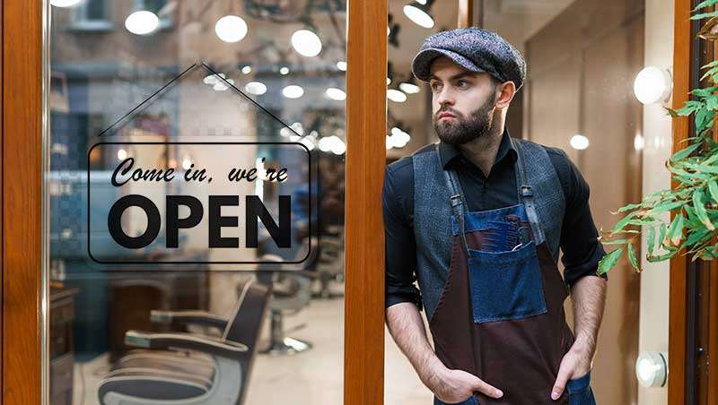 peluquero en la puerta de su salón con un cartel de open